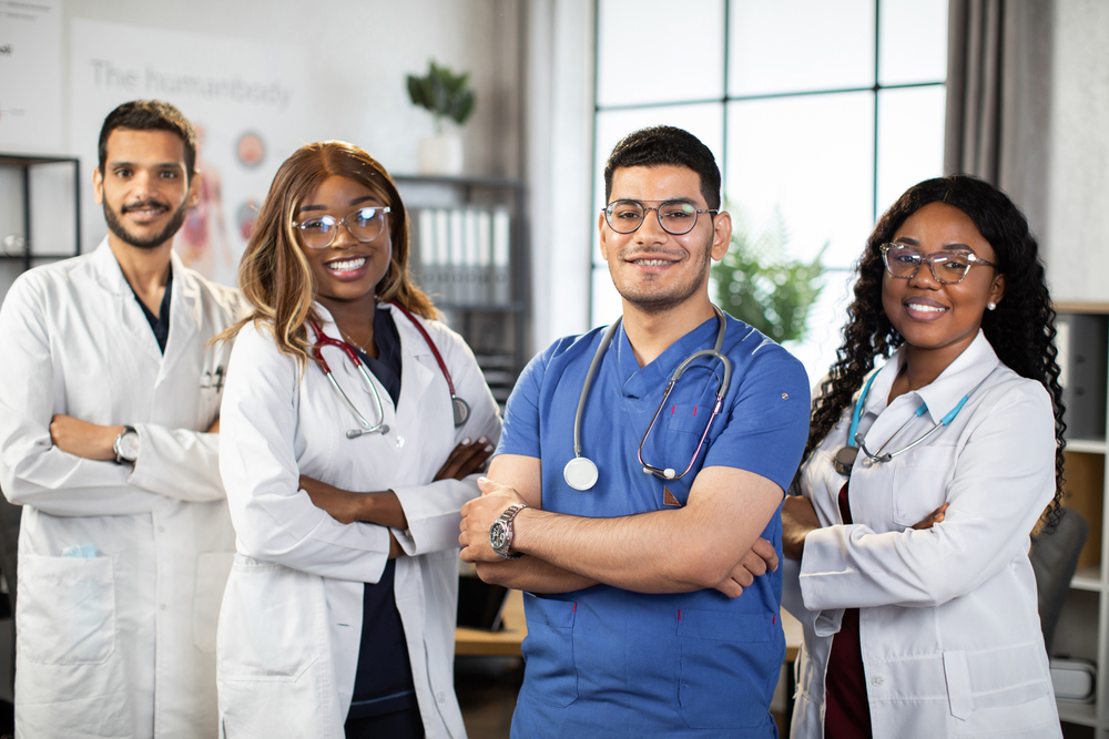 Portrait of smiling multiethnic medical team standing in modern clinic room with arms crossed. Group of pleasant confident skilled multiracial physicians posing indoors with arms folded